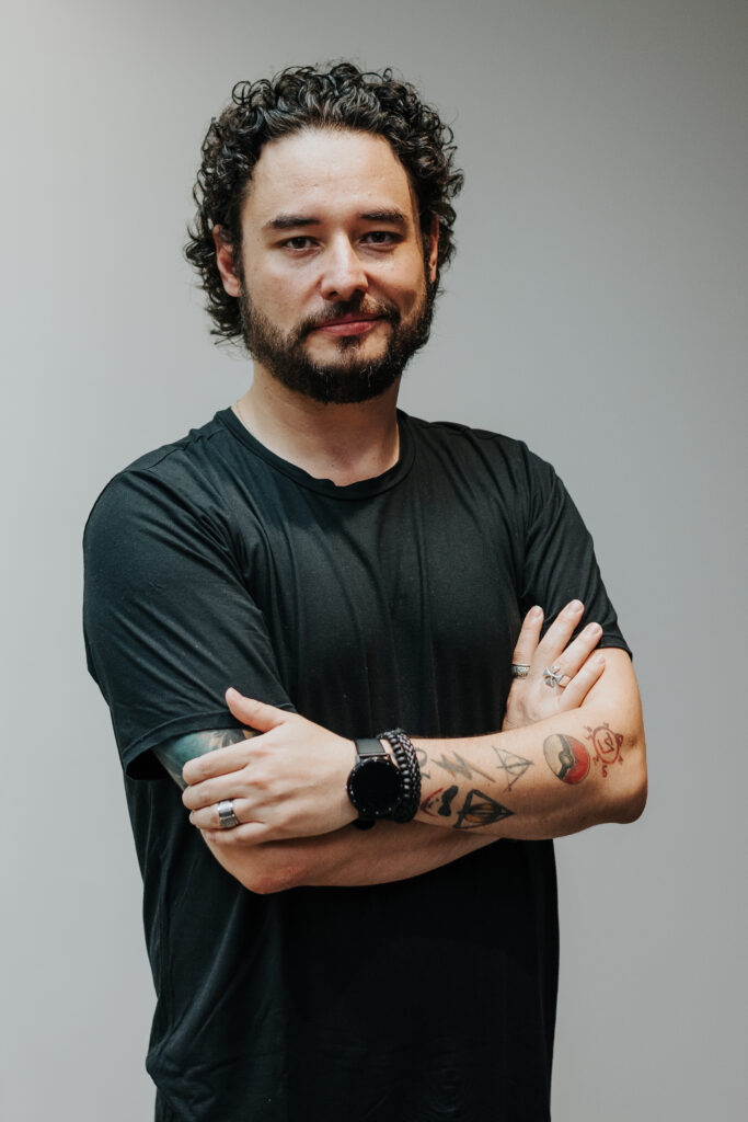 Sobre Dennis Mono Dennis Mono, mentor de líderes e times de tecnologia, de braços cruzados, expressão confiante, tatuagens visíveis.